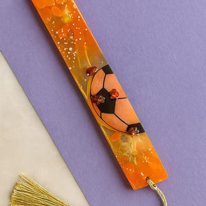 Puede incluir: Marcapáginas rectangular con diseño de balón de fútbol. El marcapáginas tiene un fondo naranja y amarillo con una borla dorada. El balón de fútbol es naranja y negro, con elementos decorativos.