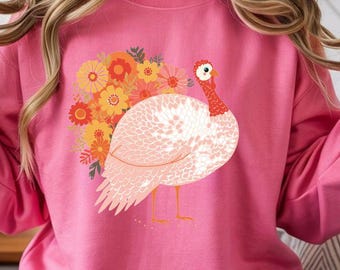 May include: Pink sweatshirt with a turkey design and floral elements. The turkey is white and pink with a red neck and yellow legs. The flowers are orange and yellow.