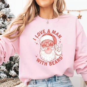 May include: Pink sweatshirt with the text "I LOVE A MAN WITH BEARD" and a graphic of Santa Claus wearing sunglasses and blowing a bubble. Santa is wearing a red hat and has a white beard. The sweatshirt is a light pink color.