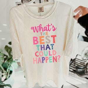 May include: Cream-colored t-shirt with the phrase "What's the BEST THAT COULD HAPPEN?" in colorful, playful lettering. The shirt is hanging on a white hanger, with a hand partially visible on the right side.
