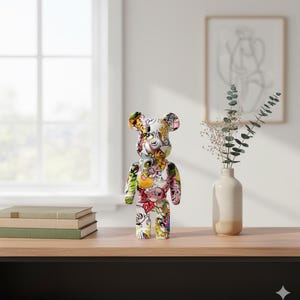 Op de afbeelding: Een kleurrijk berenbeeldje met een witte basis en diverse cartoonafbeeldingen. De beer staat op een houten oppervlak, met een raam en kunstwerk op de achtergrond. Het beeldje is een uniek decoratief item.