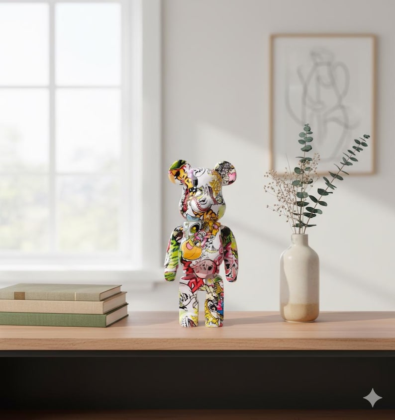 Op de afbeelding: Een kleurrijk berenbeeldje met een witte basis en diverse cartoonafbeeldingen. De beer staat op een houten oppervlak, met een raam en kunstwerk op de achtergrond. Het beeldje is een uniek decoratief item.
