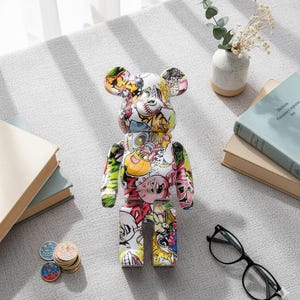 Op de afbeelding: Kleurrijke beerfiguur met diverse cartoonafbeeldingen. De beer is wit met een collage van afbeeldingen in rood, geel, groen en roze. Op een grijze ondergrond met boeken, munten, een bril en een vaasje.