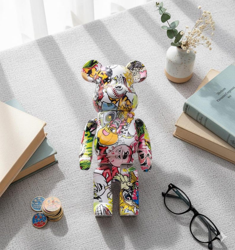 Op de afbeelding: Kleurrijke beerfiguur met diverse cartoonafbeeldingen. De beer is wit met een collage van afbeeldingen in rood, geel, groen en roze. Op een grijze ondergrond met boeken, munten, een bril en een vaasje.