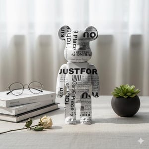 May include: A white and black decorative bear figurine with text. The bear-shaped sculpture has a newspaper print design and the words "JUSTFOR" are visible. A small succulent in a black pot, a stack of books, and round glasses are also in the image.