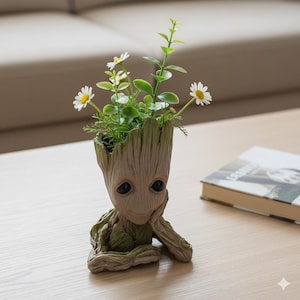 Può includere: Un vaso a forma di Groot, con un volto sorridente, pieno di margherite bianche e fogliame verde. Il vaso è marrone chiaro e poggia su una superficie di legno.