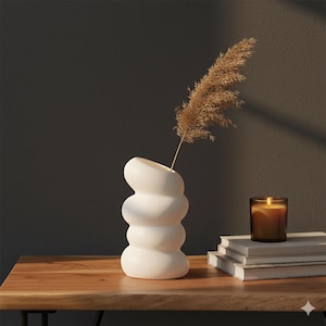 Peut inclure: Un vase blanc sculptural au design empilé unique contient une tige de plante séchée et plumeuse. Une bougie allumée dans un pot en verre est posée sur une pile de livres. Les objets sont disposés sur une table en bois sur fond sombre.
