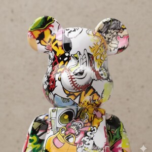 Op de afbeelding: Een witte beerfiguur met een kleurrijk graffiti-achtig ontwerp. Het ontwerp bevat honkbalsteken, cartoonfiguren en abstracte vormen in rood, geel, zwart en groen. De beer heeft grote oren en een blokkerig lichaam.
