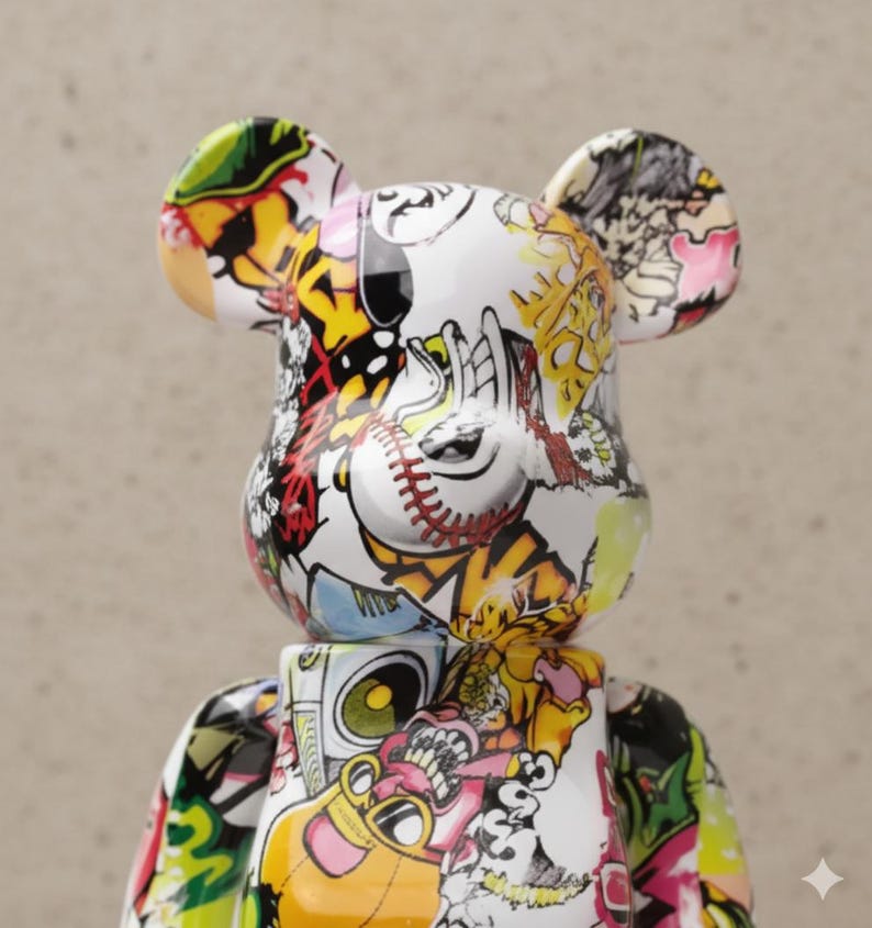 Op de afbeelding: Een witte beerfiguur met een kleurrijk graffiti-achtig ontwerp. Het ontwerp bevat honkbalsteken, cartoonfiguren en abstracte vormen in rood, geel, zwart en groen. De beer heeft grote oren en een blokkerig lichaam.