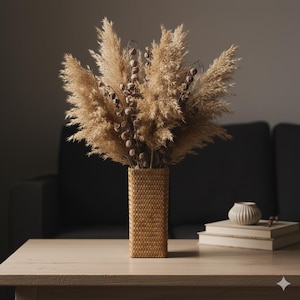 Puede incluir: Un arreglo decorativo de hierba de la pampa seca y vainas de semillas en un jarrón rectangular tejido. El jarrón es de color beige y el arreglo está sobre una mesa de madera clara. Un pequeño jarrón blanco y libros en el fondo.