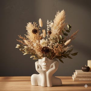 Puede incluir: Un jarrón de cerámica blanco con forma de rostro, que contiene un ramo de flores secas. El arreglo incluye hierba de la pampa, dalias marrones y varias hojas y tallos secos. El jarrón está sobre una superficie de madera.