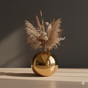 Puede incluir: Un jarrón dorado esférico con un acabado pulido que refleja el entorno. Contiene un ramo de flores y hierbas secas en tonos beige y marrón, creando una exhibición natural y elegante. El arreglo se encuentra sobre una superficie de madera clara.