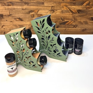 May include: Two sage green tiered spice racks featuring intricate laser-cut olive branch patterns on the sides. Each rack holds multiple cylindrical spice jars with black lids. Two additional black-lidded jars labeled Kinders, Berberine, and Biotin sit nearby.