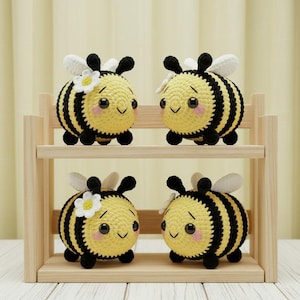 Può includere: Quattro api all'uncinetto fatte a mano con corpi a strisce gialle e nere, zampe nere e ali bianche. Due api hanno un fiore bianco sulla testa. Le api sono esposte su una mensola di legno chiaro.