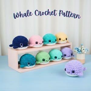 May include: A display of colorful, crocheted whale toys. The whales are in various colors, including blue, pink, green, yellow, and purple, and are arranged on a light pink shelf. The text "Whale Crochet Pattern" is above the whales.