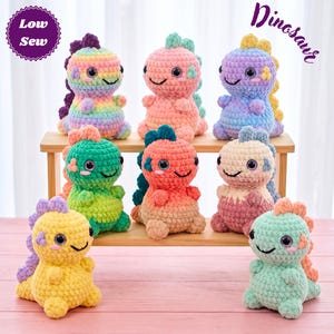 May include: Eight small, plush, crocheted dinosaur toys in various colors including rainbow, pink, blue, green, orange, cream, yellow, and mint. Each dinosaur features large dark eyes, a stitched smile, and a row of spikes along its back.