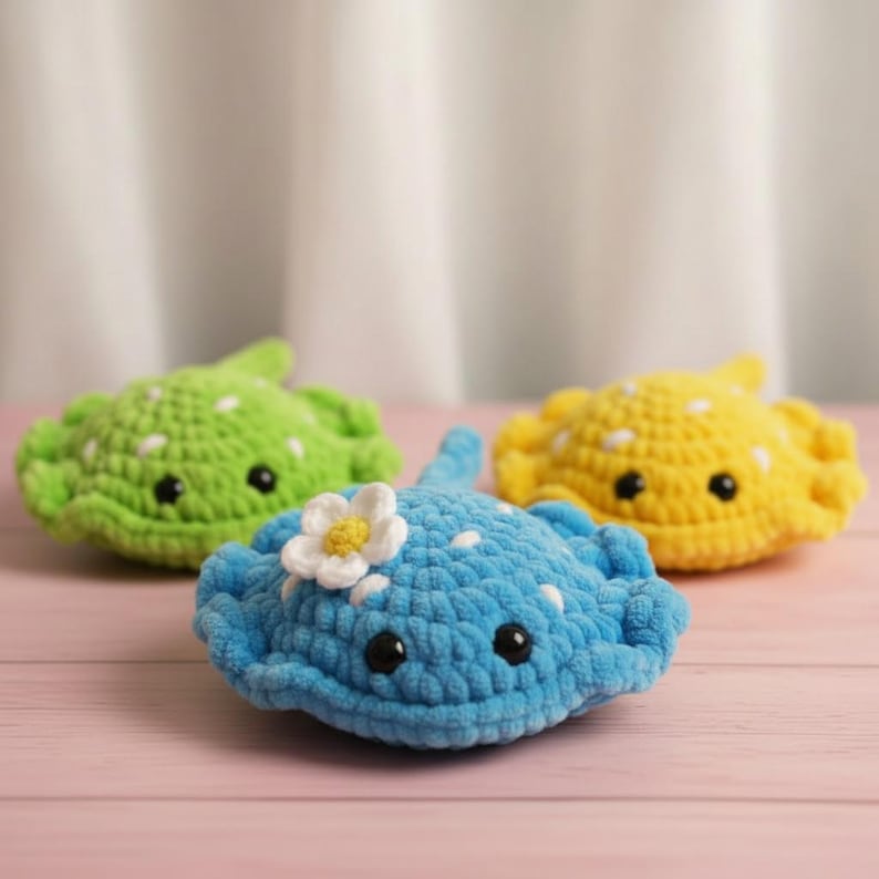 May include: Three crocheted stingrays in shades of green, blue, and yellow. The blue stingray has a white flower accent. Each has black eyes and white polka dots. The toys are arranged on a pink wooden surface.