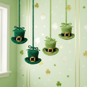 May include: Four green crochet leprechaun hats with black bands and gold buckles are suspended from green strings. The hats are hanging in front of a light green wall decorated with shamrock shapes. The hats are a festive decoration.