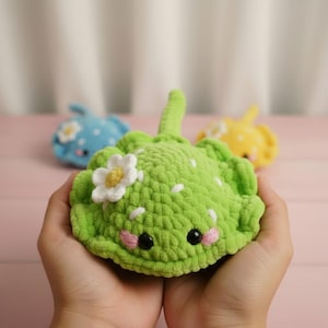 May include: A handmade green crochet stingray toy with a white flower and small white dots. The toy has black eyes and pink cheeks. Two other stingray toys are in the background, one blue and one yellow.