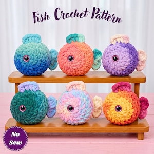 Könnte beinhalten: Sechs runde, gehäkelte Plüschfische auf einem Holzständer. Der Text 'Fish Crochet Pattern' steht oben und ein kreisförmiges Abzeichen mit dem Text 'No Sew' befindet sich unten links. Jeder Fisch hat eine weiche, strukturierte Garnoberfläche.