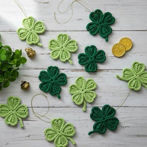 Puede incluir: Tréboles hechos a mano de ganchillo en varios tonos de verde, algunos con cuerdas para colgar, están dispuestos sobre una superficie clara de vetas de madera. También hay monedas de oro y un pequeño pote de oro, evocando el Día de San Patricio.