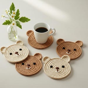 May include: A collection of handmade crochet bear coasters in shades of beige and brown. Each coaster features a bear face design with black eyes and a brown nose. A white mug filled with coffee sits on one of the coasters.