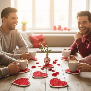 K&ouml;nnte beinhalten: Vier Personen sitzen lachend um einen Holztisch und halten Kaffeetassen. Rote herzf&ouml;rmige Untersetzer und Rosenbl&auml;tter schm&uuml;cken den Tisch. Eine Vase mit einer Rose steht in der Mitte, was auf ein romantisches Thema hindeutet.