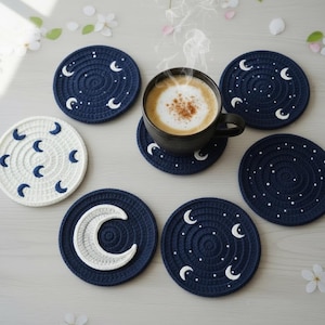 May include: A collection of handmade crochet coasters in navy blue and white. The coasters feature crescent moon and star designs. A black mug with a cappuccino sits on one of the coasters. The coasters are round and have a spiral pattern.