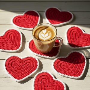 Könnte beinhalten: Eine klare Glastasse Kaffee mit Latte Art steht auf einem roten herzförmigen Untersetzer. Mehrere rote herzförmige Untersetzer mit weißem Rand umgeben die Tasse. Die Untersetzer sind aus gehäkeltem Material.