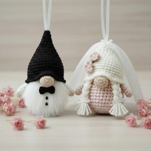 May include: Two handmade crochet gnomes, one dressed as a groom in a black hat and bow tie, the other as a bride in a white veil and hat with pink flowers. The gnomes have white beards and are decorated with pink flowers.