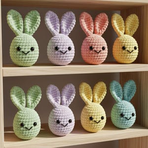 May include: A collection of eight crocheted Easter bunny decorations in various pastel colors, including green, lavender, peach, and yellow. Each bunny has a smiling face and upright ears, displayed on wooden shelves.