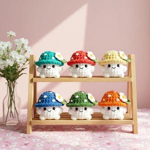 May include: Six handmade crochet mushroom figures with smiling faces and flower accents are displayed on a wooden shelf. Each mushroom has a different coloured hat: teal, red, yellow, blue, green, and orange. The shelf is on a pink floral patterned surface.