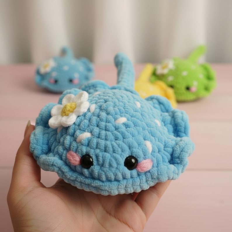 May include: A handmade, light blue crochet stingray with white spots and a white flower with a yellow center. The stingray has black eyes and pink cheeks. Two other crochet stingrays are in the background, one blue and one green.