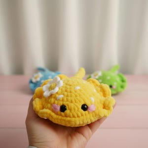 May include: A handmade yellow crochet stingray with white polka dots and a white flower accent. The stingray has black eyes and pink cheeks. Other crochet stingrays in blue and green are in the background.