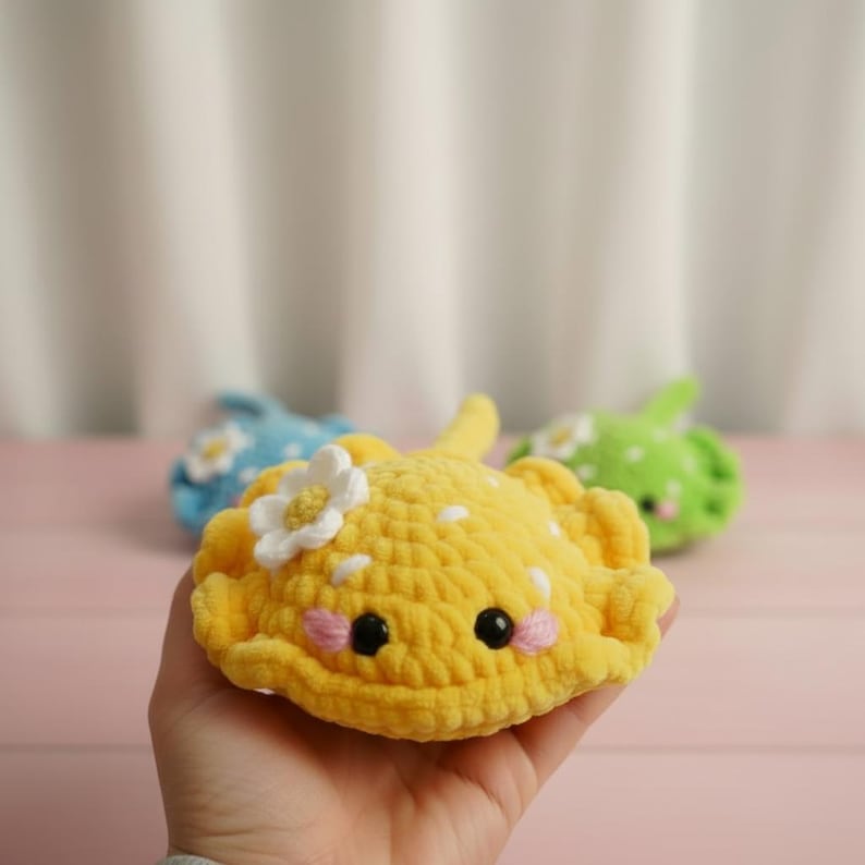 May include: A handmade yellow crochet stingray with white polka dots and a white flower accent. The stingray has black eyes and pink cheeks. Other crochet stingrays in blue and green are in the background.