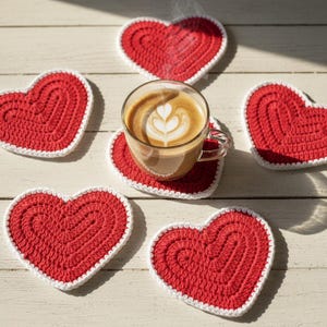 K&ouml;nnte beinhalten: Eine klare Glastasse Kaffee mit Latte Art steht auf einem roten herzf&ouml;rmigen Untersetzer. Mehrere rote herzf&ouml;rmige Untersetzer mit wei&szlig;em Rand umgeben die Tasse. Die Untersetzer sind aus geh&auml;keltem Material.