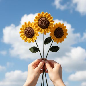 May include: A bouquet of three handmade crochet sunflowers. The flowers have yellow petals and brown centers, with green leaves and stems. The bouquet is held against a blue sky with white clouds.