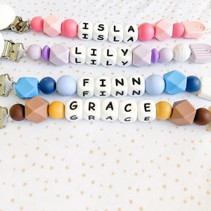 Puede incluir: Cuatro clips de chupete personalizados con los nombres Isla, Lily, Finn y Grace. Cada clip presenta una variedad de cuentas coloridas, incluyendo formas geométricas y cuentas redondas, y un clip de metal. Los clips de chupete son en tonos rosa, morado, azul, marrón y amarillo.