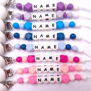 Puede incluir: Una colección de ocho clips para chupetes con broches de plástico blanco redondos y cordones blancos. Cada clip incluye una secuencia de cuentas de silicona de colores en tonos morado, azul y rosa, intercaladas con cuentas cuadradas blancas que deletrean NAME.