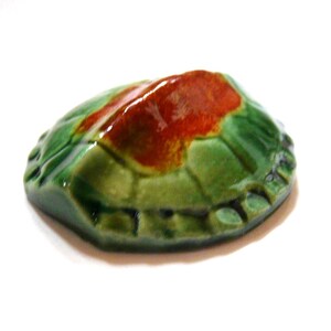 May include: A ceramic turtle figurine with a glossy finish. The turtle's shell is primarily green with a reddish-brown patch on top. The edges of the shell have a textured, scalloped design. The figurine is set against a white background.