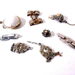 May include: A collection of vintage jewelry components, including clasps, a bow-shaped piece, and decorative elements with rhinestones and pearls. The pieces are in silver, gold, and cream colors, with intricate detailing.
