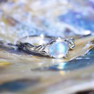 May include: A silver ring with a large, round, iridescent moonstone. The ring has a delicate, leaf-like design on the band. The ring is displayed on a multi-colored, iridescent surface, creating a dreamy aesthetic.
