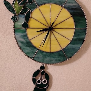 May include: A stained glass clock featuring a frog theme. The clock face is yellow and green, with a frog perched on top and another hanging below. The clock hands are black.