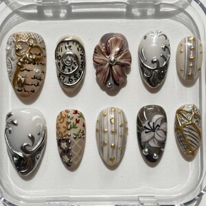 May include: A collection of ten artificial nails presented in a clear case. The nails showcase diverse designs, such as floral motifs, clock faces, and decorative elements in gold, silver, and brown. The nails are oval-shaped.
