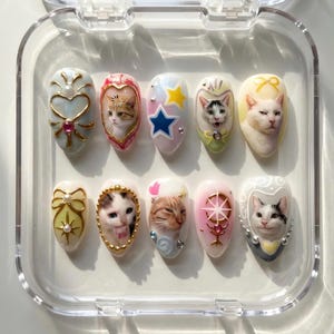 May include: A set of ten artificial nails with various cat-themed designs. Each nail features a different cat portrait or decorative element, including hearts, stars, and bows. The nails are displayed in a clear plastic case.