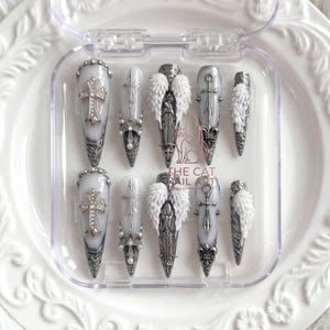 May include: A set of ten stiletto-shaped press-on nails in a clear plastic case. The nails feature a white base with silver metallic accents, including cross charms, small white pearls, and textured white angel wing embellishments. The text THE CAT NAIL ART is printed in the centre.