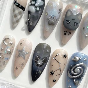 May include: A collection of almond-shaped false nails with various designs. The designs include a plaid pattern, paw prints, a cat face, stars, constellations, and a galaxy. The nails are in a clear plastic case.