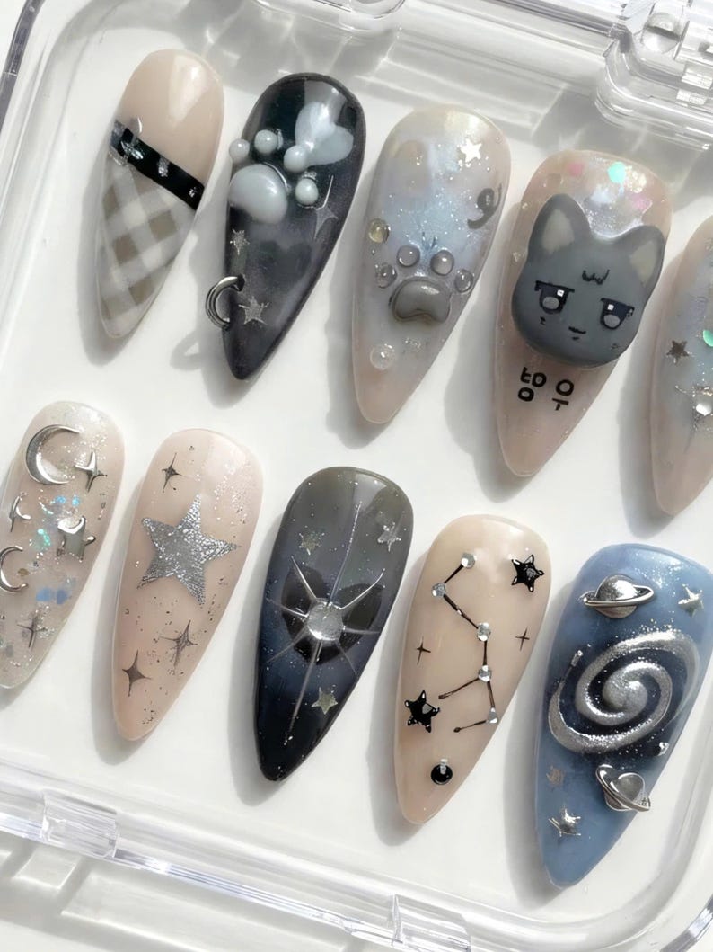 May include: A collection of almond-shaped false nails with various designs. The designs include a plaid pattern, paw prints, a cat face, stars, constellations, and a galaxy. The nails are in a clear plastic case.