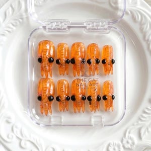 May include: A set of ten orange, translucent, shrimp-shaped press-on nails with black bead eyes, arranged in two rows inside a clear plastic storage case. The case features the text THE CAT NAIL ART PRESS ON NAILS printed in the centre.