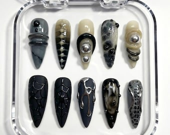 May include: A set of ten artificial nails in a clear plastic case. The nails feature various designs, including spiderwebs, metallic drips, and textured patterns. Colors include black, gray, and beige, with silver accents. The nails are pointed and have a glossy finish.
